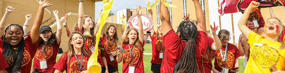 photo of about a dozen Ƶ students wearing matching marron and gold shirts cheer with their arms raised. With streamers and flags flying, the students exude high energy on a grassy lawn with yellow brick buildings in the background.